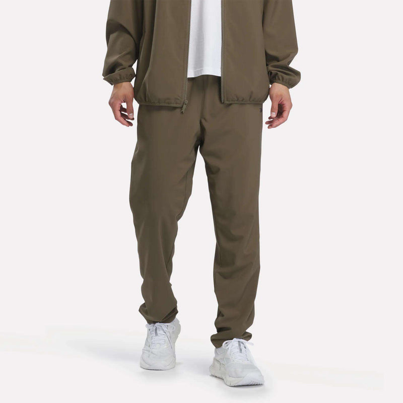 A person wearing Reebok Men’s Woven Training Pants in army green, paired with a white shirt and white sneakers, standing against a plain white background.