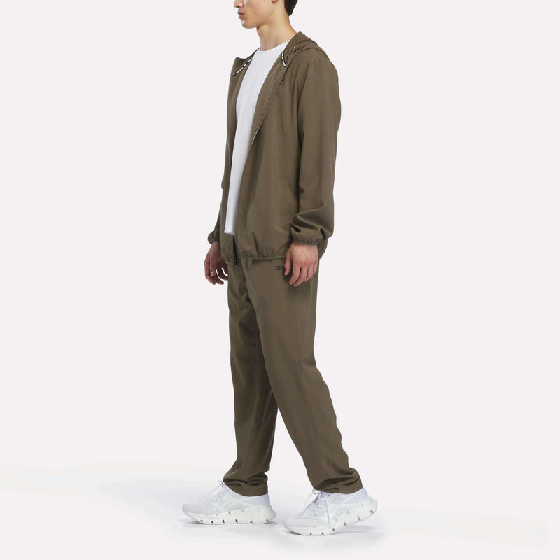 A person wearing Reebok Men’s Woven Training Pants in brown, paired with a white t-shirt and white sneakers, stands against a plain white background.