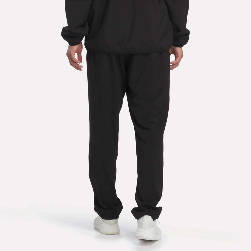Person standing with their back to the camera, wearing Reebok Men’s Woven Training Pants in black and white sneakers, against a plain white background.