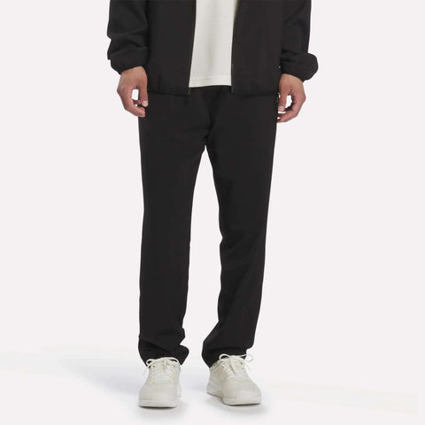 A person is shown from the waist down wearing Reebok Men’s Woven Training Pants in black, paired with a white shirt, a black jacket, and white sneakers, standing against a plain white background.