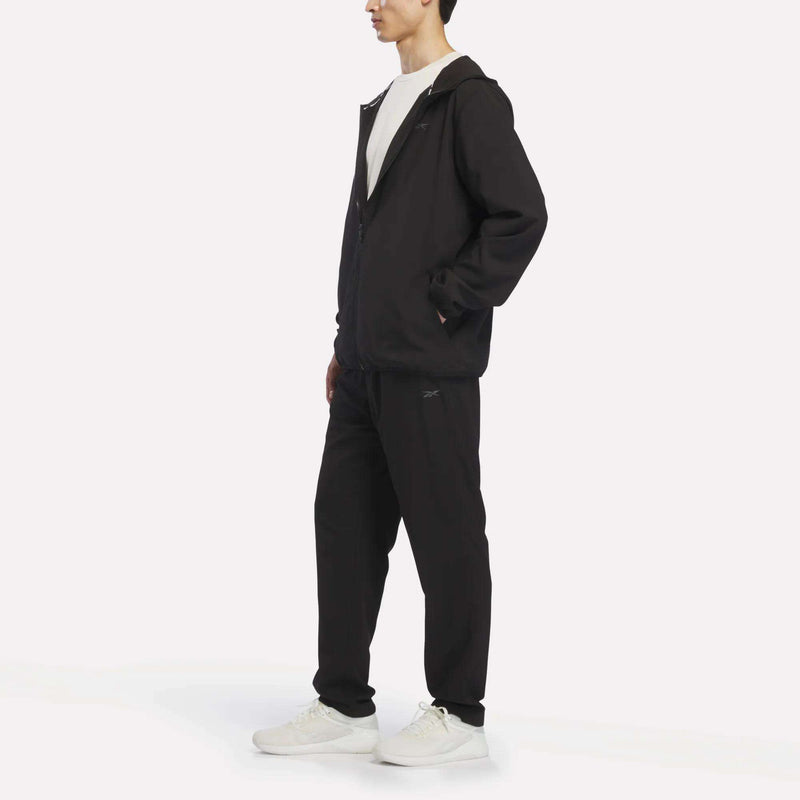 A person wears a black zip-up jacket, Reebok Men’s Woven Training Pants, a white t-shirt, and white sneakers, standing against a plain white background.
