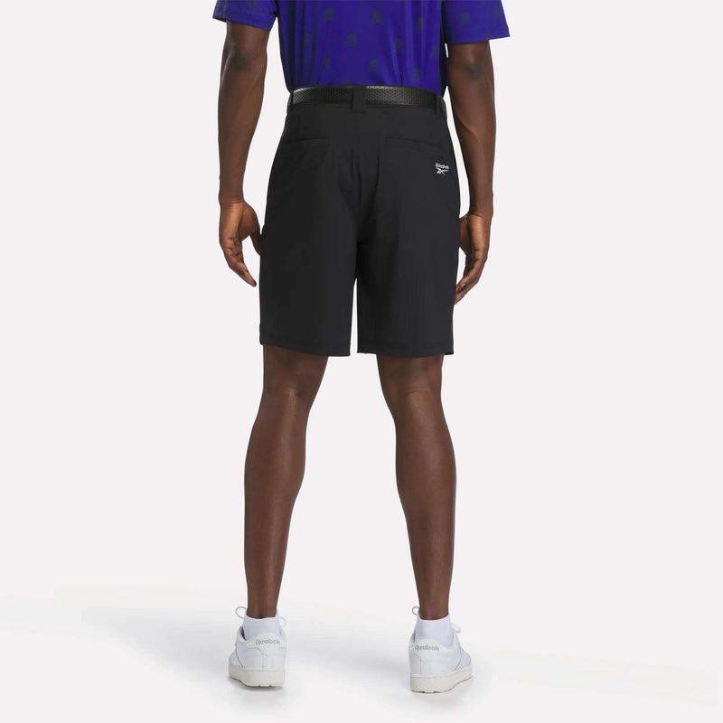 A man stands facing away, wearing a blue shirt and Reebok Men's Pull-On Golf Shorts 8.5" in black with a belt, featuring four-way stretch for comfort and ease of movement, paired with white sneakers against a plain white background.