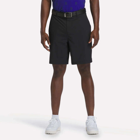 A person stands against a plain white background wearing a blue shirt and the Reebok Men's Pull-On Golf Shorts 8.5" in black, which feature four-way stretch for added comfort. The look is completed with white sneakers.