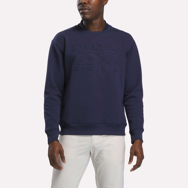 A man stands against a plain white background wearing the Reebok Men's Signature Golf Crewneck Sweatshirt in Midnight Blue, which features embossed lettering and advanced RBK-DRY technology, paired with white pants.