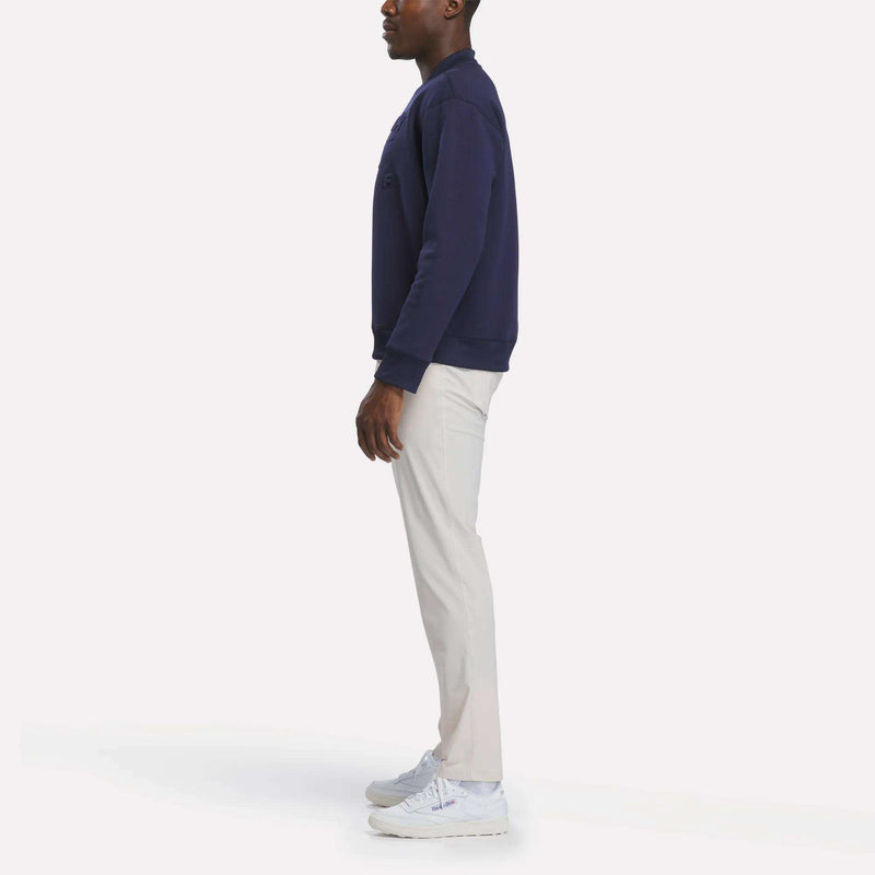 A person stands sideways against a plain white background, wearing the Reebok Men's Signature Golf Crewneck Sweatshirt in navy, paired with light-colored pants and white sneakers.
