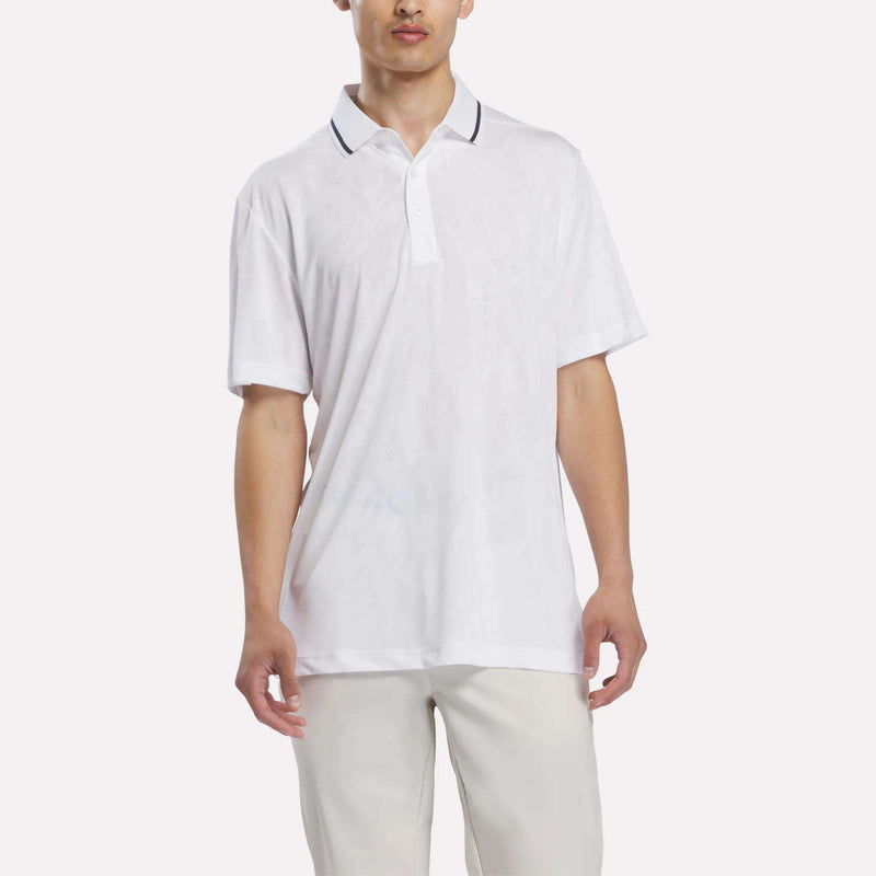 A person wearing a white Reebok Men’s Tournament Polo Shirt with short sleeves and a collar, paired with light-colored pants, stands against a plain white background.