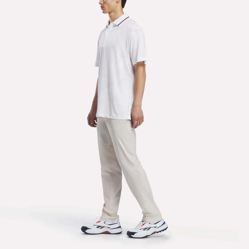 A man stands in profile wearing the Reebok Men’s Tournament Polo Shirt in white, paired with light beige pants and white sneakers with black and red accents. The background is plain white.