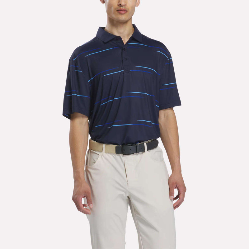 A man wearing a Reebok Men’s Tournament Polo Shirt in Vector Navy, paired with beige pants and a black belt, stands against a plain white background. This polo features a subtle tournament-inspired print for a sporty, refined look.