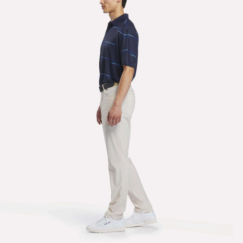 A man stands sideways wearing the Reebok Men’s Tournament Polo Shirt, featuring navy blue stripes and a subtle tournament-inspired print, paired with light beige pants, a black belt, and white sneakers against a plain white background.