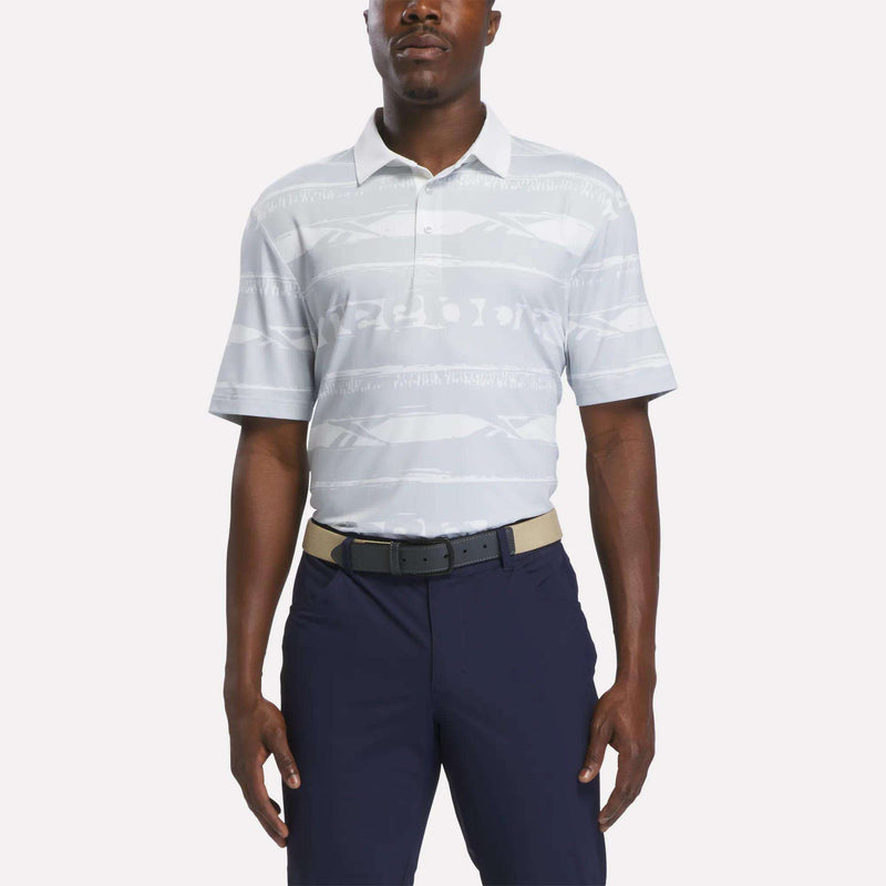 A man wears the Reebok Men's Tournament Polo Shirt in Grey/White, styled with navy blue pants and a beige belt, standing against a plain white background.