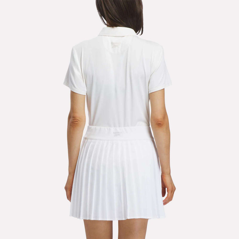 A woman stands facing away against a plain background, wearing a short-sleeved Reebok Women's Solid Core Polo Shirt in white and a white pleated skirt.
