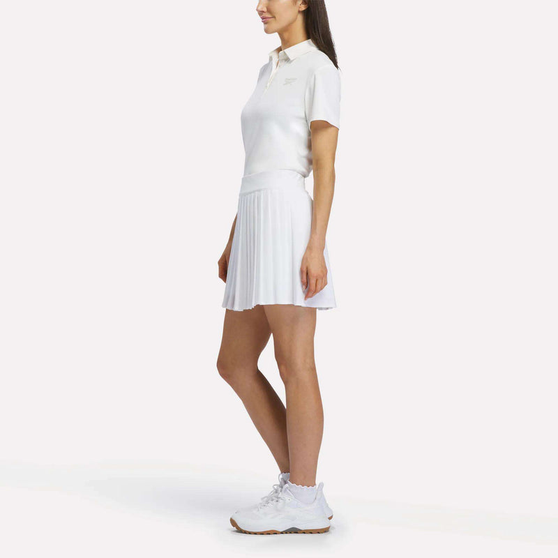 A woman stands sideways wearing the Reebok Women's Solid Core Polo Shirt in white, paired with a white pleated tennis skirt and white sneakers against a plain white background.