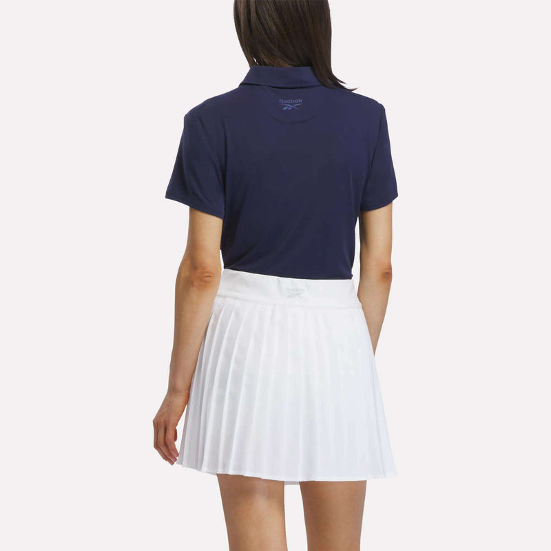 A woman stands with her back to the camera, wearing a dark blue Reebok Women's Solid Core Polo Shirt and a white pleated skirt. Her straight, shoulder-length hair contrasts with the plain, light-colored background.