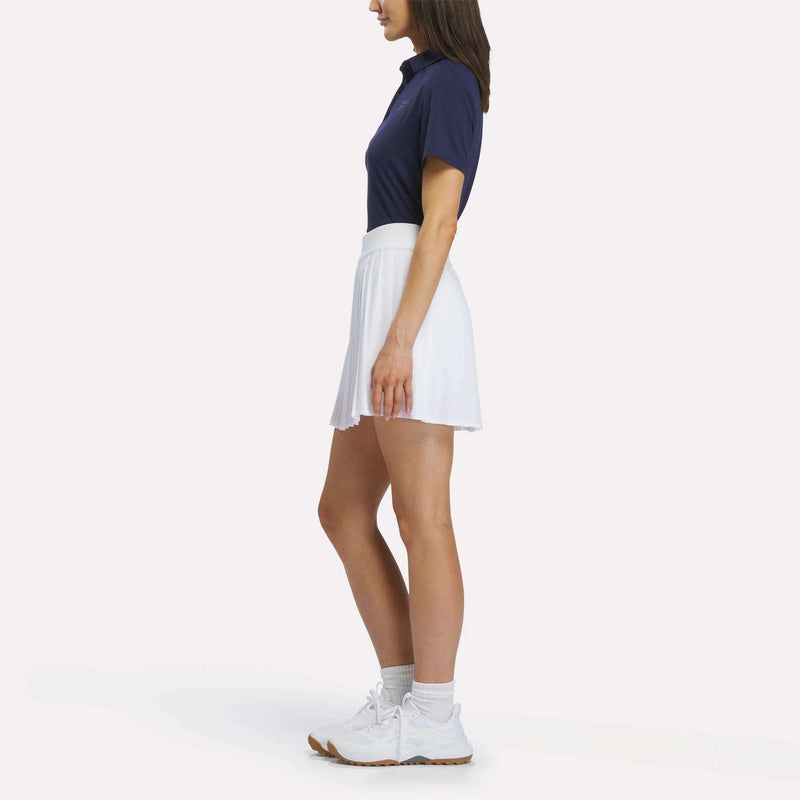 A woman stands sideways against a plain white background, wearing the Reebok Women's Solid Core Polo Shirt in navy, paired with a white pleated skirt, white socks, and athletic shoes.