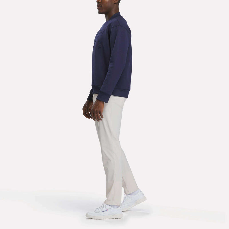 A person stands sideways against a plain white background wearing a navy sweatshirt, white sneakers, and Reebok Men's Favorite 5-Pocket Relaxed Fit Golf Pants in a wrinkle-resistant fabric.