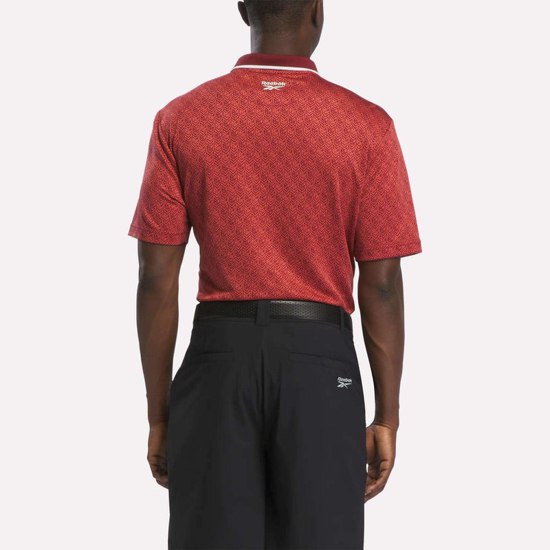A man is shown from the back, standing against a plain white background, wearing a Reebok Men's Jacquard Polo Shirt in a red pattern and black pants.