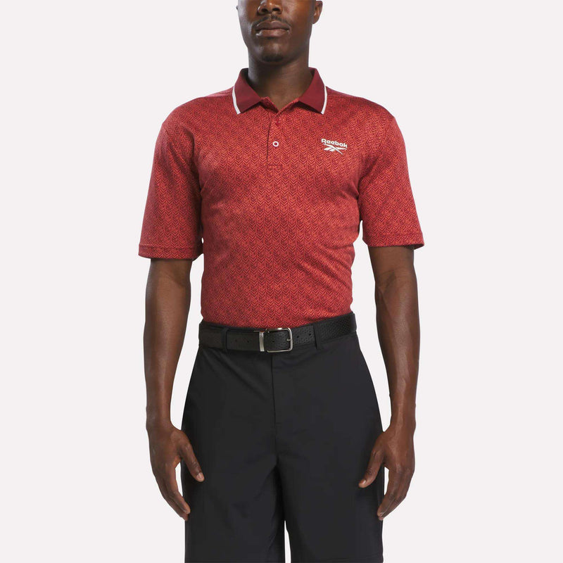 A man stands facing forward against a white background wearing a brick red/chalk Men's Jacquard Polo Shirt from Reebok, featuring a white logo on the chest, paired with a black belt and black shorts.