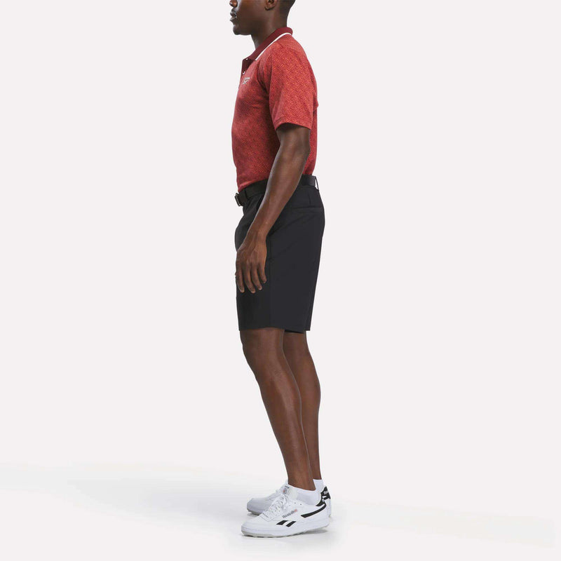 A man stands in profile wearing a Reebok Men's Jacquard Polo Shirt in red, paired with black shorts, white sneakers, and a black belt against a plain white background.