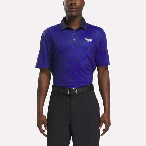 A man wearing a Reebok Men's Crushers Polo Shirt in Vector Navy and Trophy Gold, featuring a dark blue design, stands with his arms relaxed at his sides against a plain background.