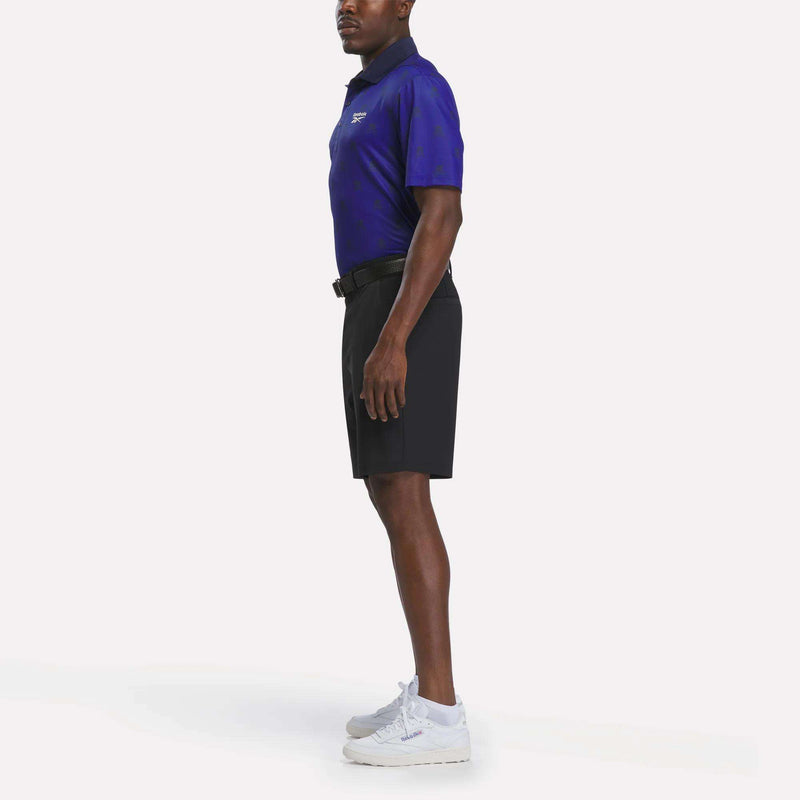 A man stands in profile wearing a dark blue Reebok Men's Crushers Polo Shirt, black shorts, white sneakers, and a black belt against a plain white background.