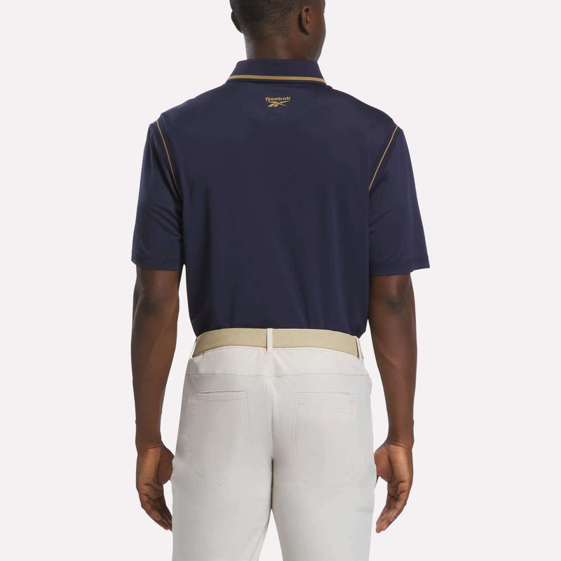 A man stands facing away, wearing a navy Men's Varsity Polo Shirt by Reebok with gold trim and light beige pants. The comfortable golf polo features a small logo under the collar. He pairs it with a beige belt, his arms relaxed at his sides.