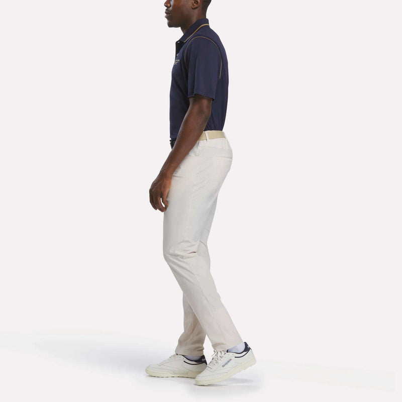A man stands sideways against a plain white background, wearing a Reebok Men's Varsity Polo Shirt made with UPF-rated fabric, beige pants, white sneakers, and a beige belt.