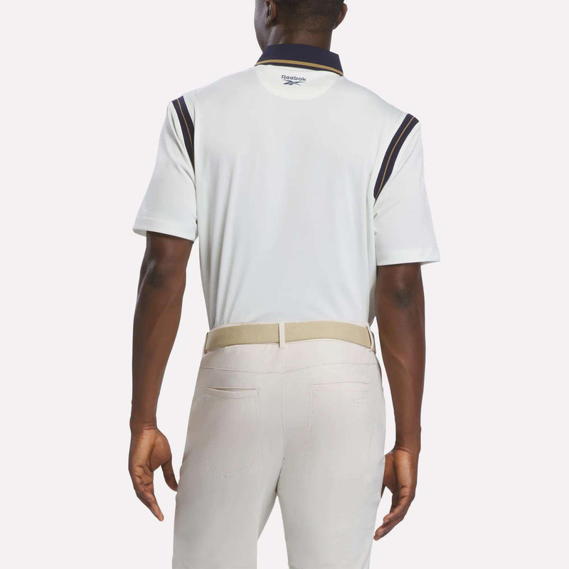 A man is shown from the back wearing a Men's Varsity Polo Shirt by Reebok, featuring RBK-DRY fabric with navy accents. He pairs it with light beige pants and a light-colored belt. The background is plain white.