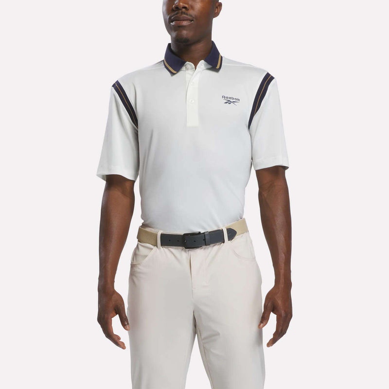 A man wearing a chalk-colored Men's Varsity Polo Shirt by Reebok, crafted from moisture-wicking RBK-DRY fabric and featuring navy accents, along with beige pants and a black belt, stands with his arms relaxed at his sides against a plain background.