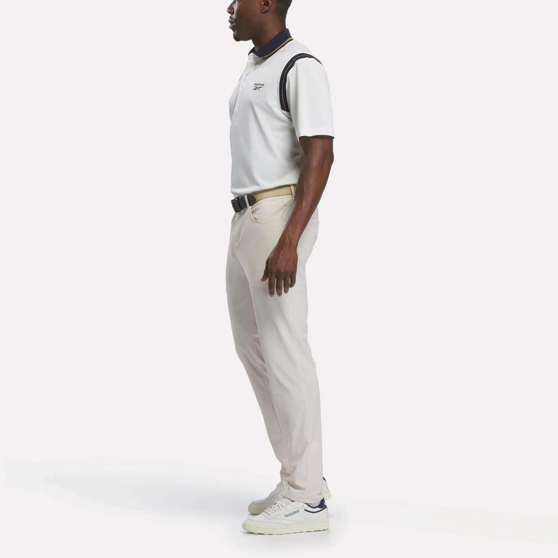 A man stands in profile wearing a crisp Reebok Men's Varsity Polo Shirt, paired with light beige pants and white sneakers. His hands are relaxed by his sides against a plain white background.