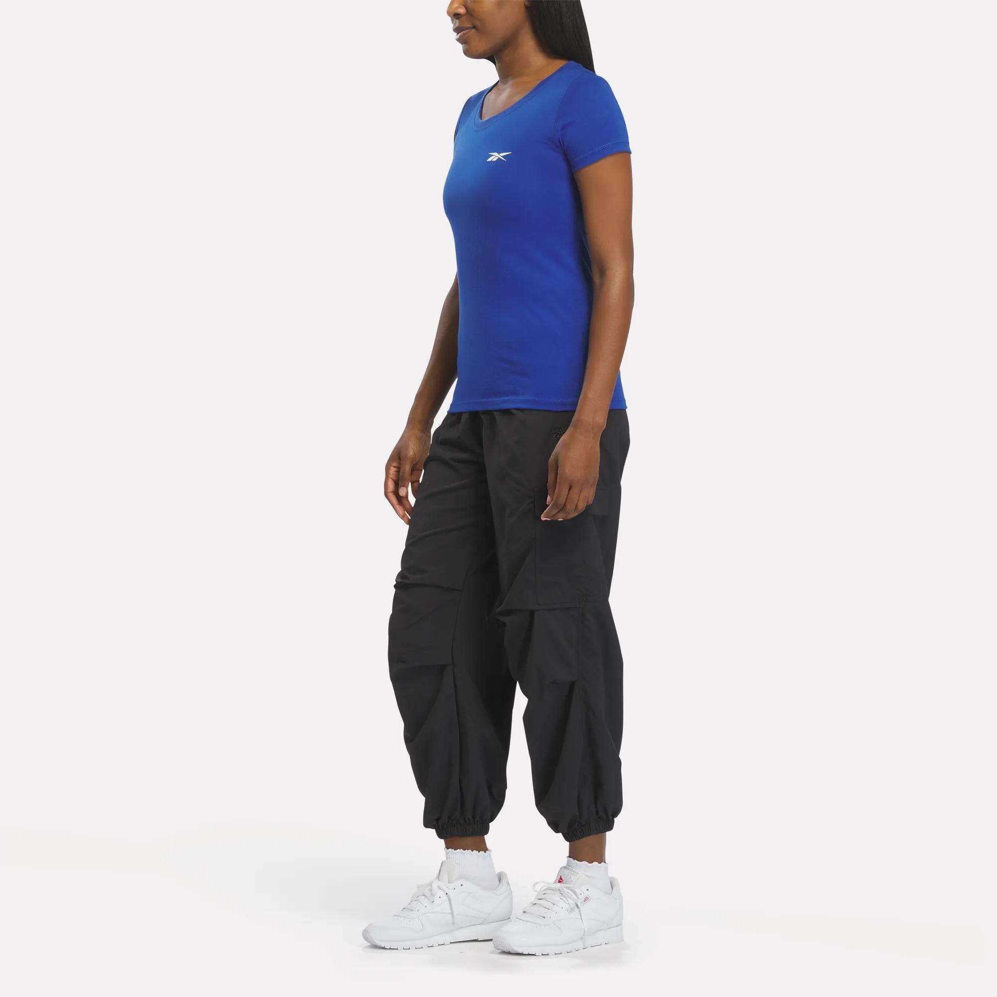 A woman faces left against a plain white background, wearing a fitted blue Reebok Women's T-Shirt, black cargo jogger pants, and white athletic sneakers.