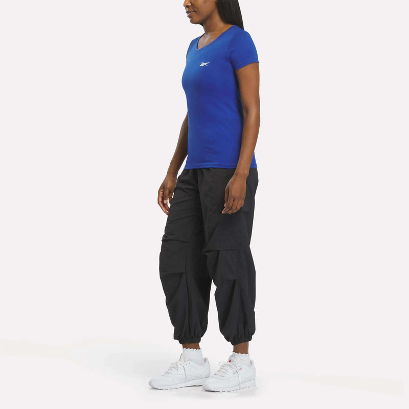 A woman faces left against a plain white background, wearing a fitted blue Reebok Women's T-Shirt, black cargo jogger pants, and white athletic sneakers.