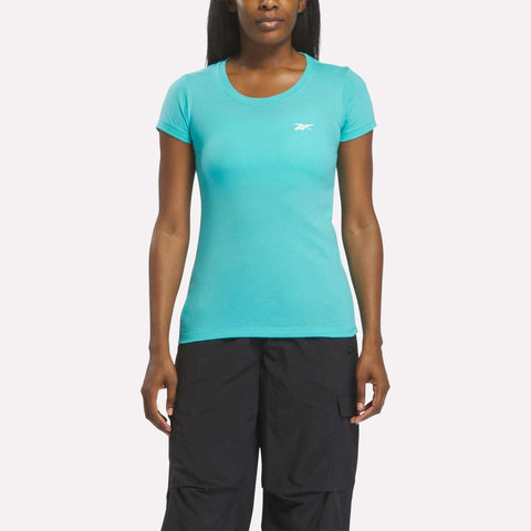 A woman in a fitted, short-sleeve Reebok Women's T-Shirt in Thati Blue and black pants stands facing forward against a plain white background.