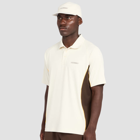 A man wearing the Reebok x Manors Men’s Polo in light beige, featuring brown side panels, along with a matching beige cap, stands against a plain light gray background.