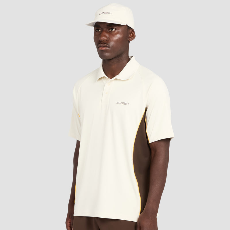 A man wearing the Reebok x Manors Men’s Polo in light beige, featuring brown side panels, along with a matching beige cap, stands against a plain light gray background.