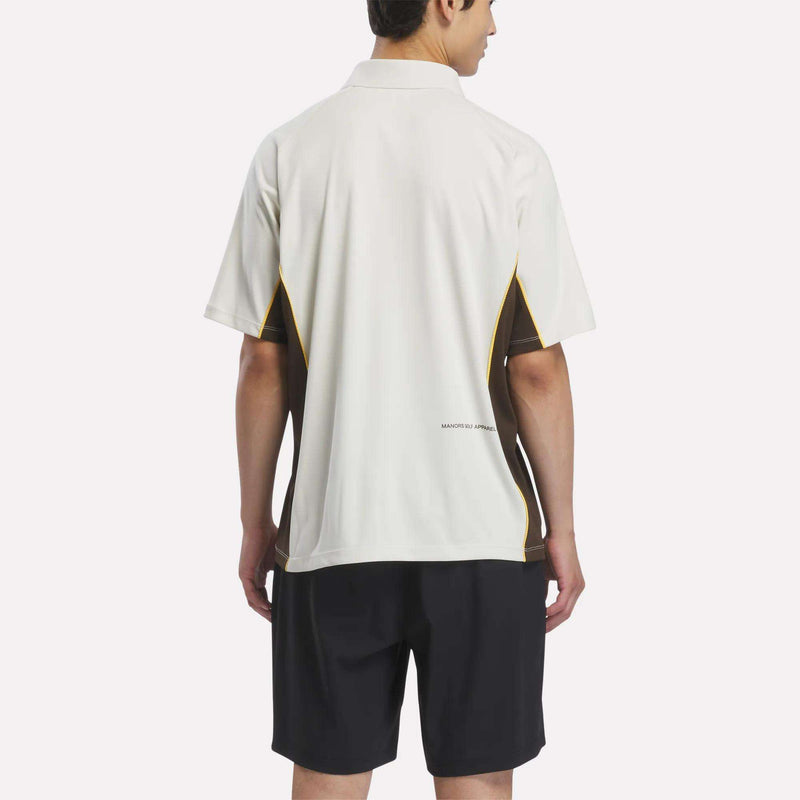 A person stands facing away, wearing a Reebok x Manors Men’s Polo in white and black with yellow accents, paired with black shorts, against a plain light background.