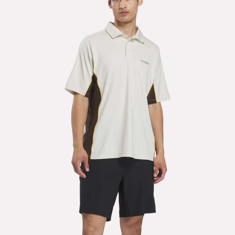A man stands facing forward against a plain white background, wearing the Reebok x Manors Men’s Polo by Reebok, featuring white short sleeves with black and yellow side panels, paired with black shorts.