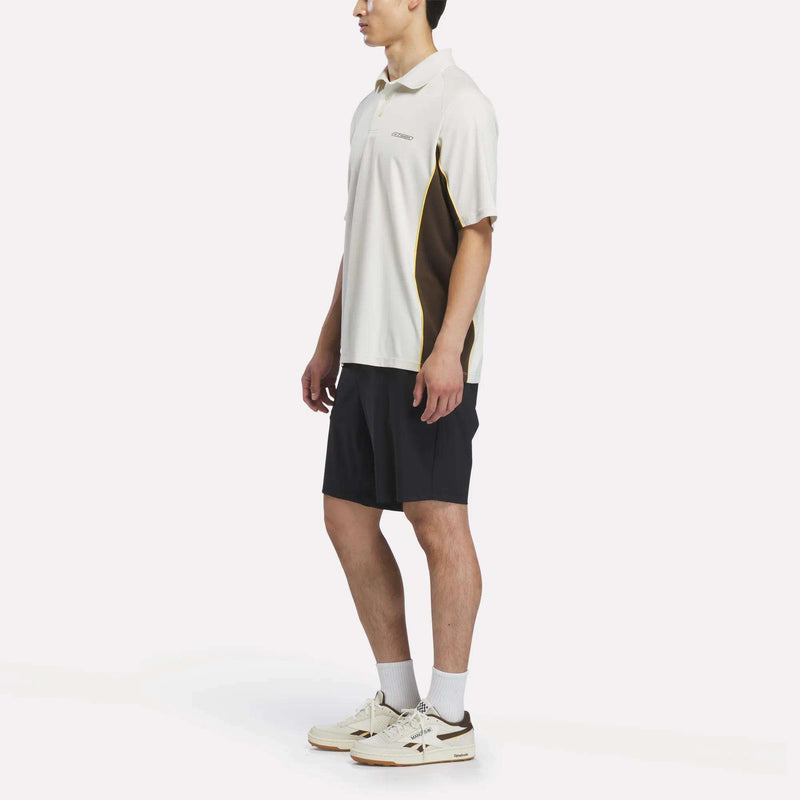 A person stands in profile against a plain white background wearing the Reebok x Manors Men’s Polo by Reebok, styled with black shorts, white socks, and white sneakers.