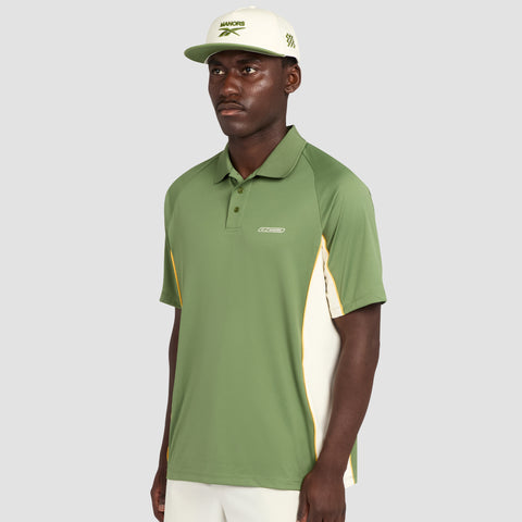 A man wearing the Reebok x Manors Men’s Polo in green and white, along with a white cap, stands against a light gray background, looking forward with a neutral expression.