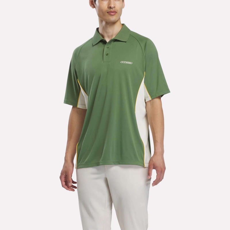 A person is wearing the Reebok x Manors Men’s Polo by Reebok, featuring green fabric with white and yellow accents, paired with white pants, while standing against a plain white background.