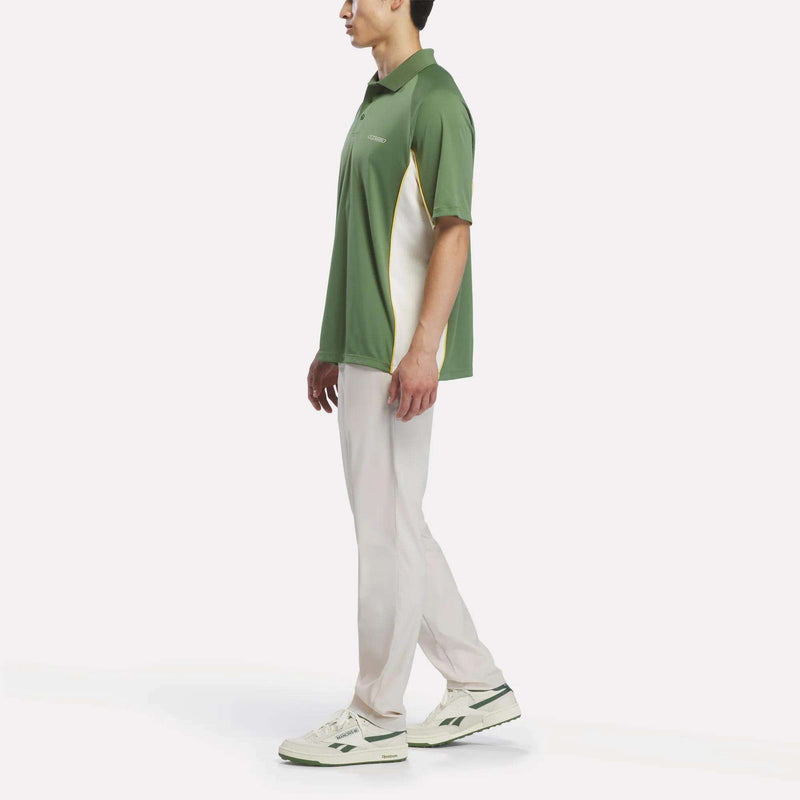 A man stands sideways against a plain white background, wearing the Reebok x Manors Men’s Polo in green with white accents, paired with light-colored pants and white sneakers featuring green details.