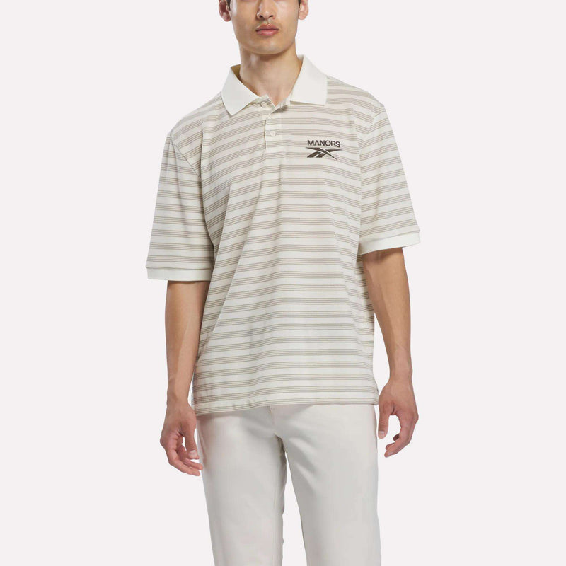 A man wears the Reebok x Manors Men’s Pique Polo, a beige and white striped golf shirt from Reebok, featuring MANORS and a logo on the left chest. He pairs this men’s golf polo with white pants while standing against a plain light background.