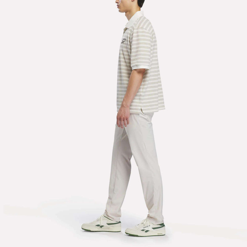 A person wearing the Reebok x Manors Men’s Pique Polo in beige and white stripes, light pants, and white sneakers stands facing left against a plain white background.