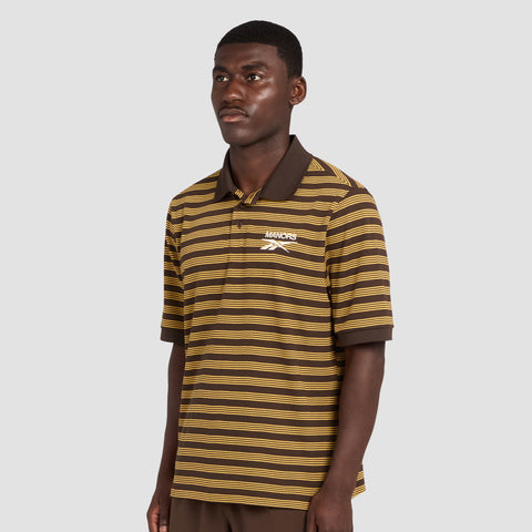 A man wearing a Reebok x Manors Men’s Pique Polo in brown and yellow stripes, featuring “Manors” and a logo on the chest, stands against a plain light gray background.