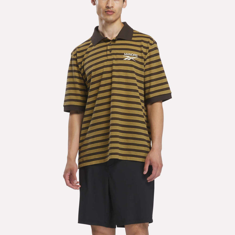 A person wearing the Reebok x Manors Men’s Pique Polo in Espresso/Gorse and black shorts stands against a plain white background.