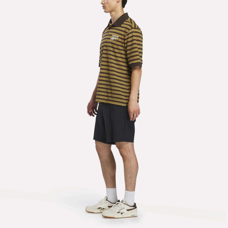 A person wearing a Reebok x Manors Men’s Pique Polo in brown and black stripes, paired with black shorts, white socks, and white sneakers stands against a plain white background.