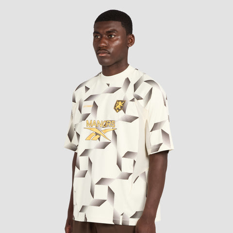 A man wears the Reebok x Manors Men’s Mockneck Polo, featuring a cream base with a geometric black and gray pattern, a lion crest, and MANORS alongside Reebok x Manors text on the front. Crafted from performance stretch fabric by Reebok, he stands against a plain gray background.