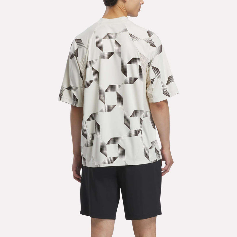 A person is standing facing away, wearing the Reebok x Manors Men’s Mockneck Polo—a short-sleeve white shirt with a black geometric pattern crafted from performance stretch fabric—paired with black shorts against a plain background.