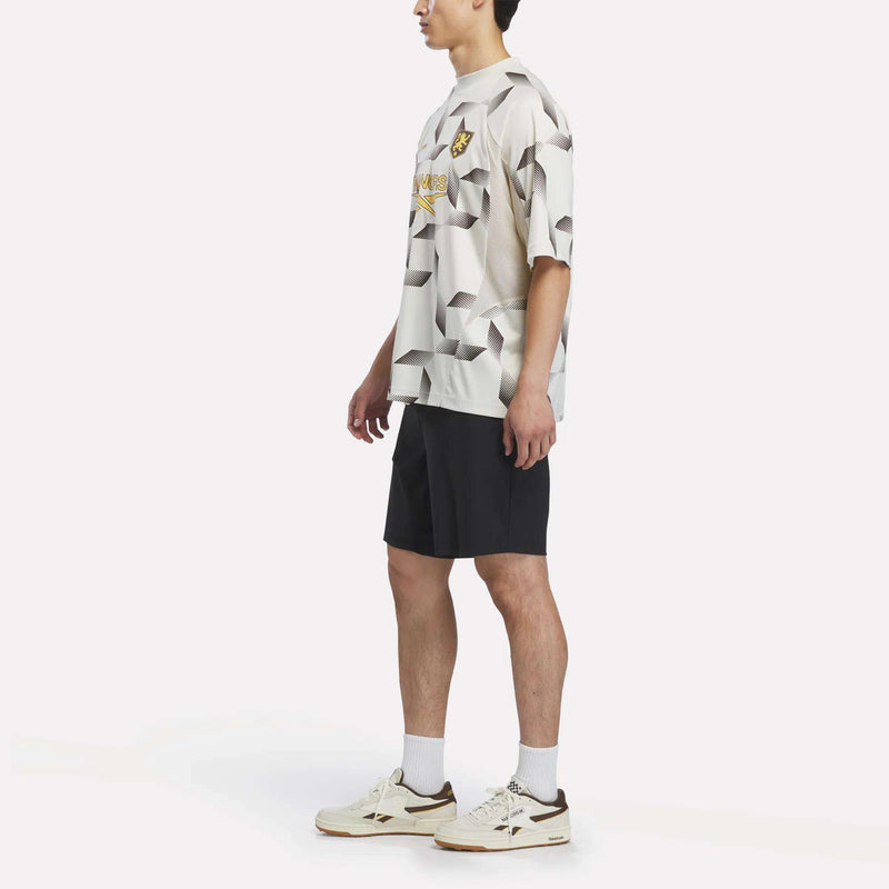 A person stands sideways against a plain white background, wearing the Reebok x Manors Men’s Mockneck Polo by Reebok in a beige patterned performance stretch fabric, paired with black shorts, white socks, and Reebok x Manors white sneakers.