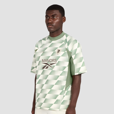 A man stands against a plain light gray background wearing the Reebok x Manors Men’s Mockneck Polo by Reebok, a green and white checkered sports jersey made from performance stretch fabric and adorned with “MANORS” and a lion crest on the front. This striking piece highlights the collaborative spirit between Reebok and Manors while offering the comfort of a men’s mockneck polo.