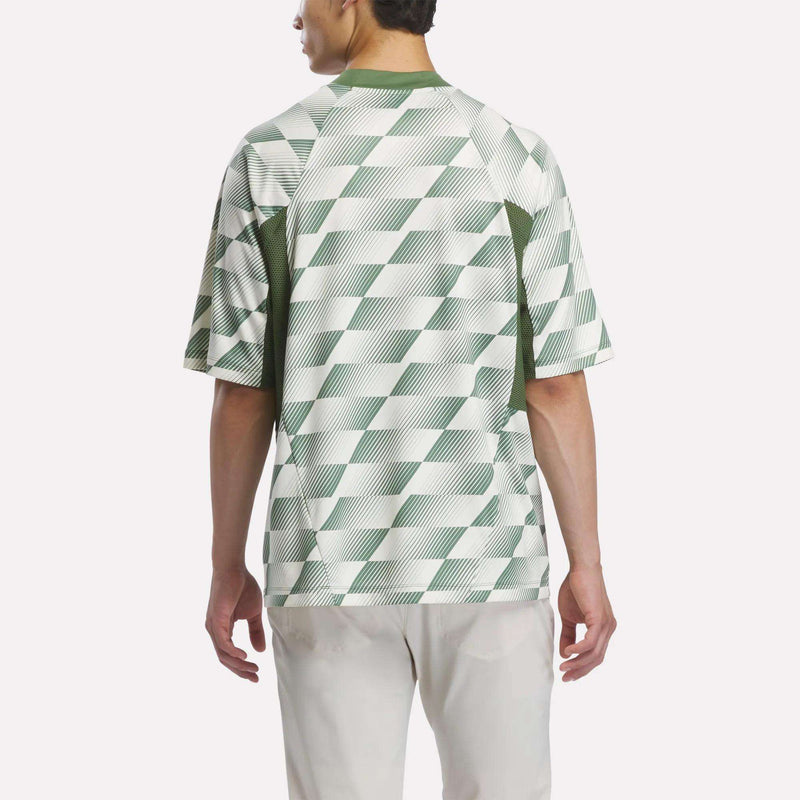 A person stands facing away, wearing the Reebok x Manors Men’s Mockneck Polo by Reebok, featuring a green and white geometric pattern and light-colored pants, crafted from performance stretch fabric, set against a plain background.
